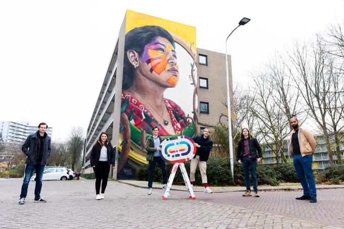 Vijf studenten staan voor een flatgebouw met een grote muurschildering erop.