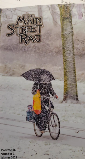 Cover of Main Street Rag winter edition with person on a bike in a snowstorm holding umbrella and shopping bags precariously past a tree on a plowed road
