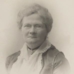 A black and white photograph of a woman dressed in old-fashioned clothing, wearing small, round glasses. She is looking towards the camera and isn't smiling. 