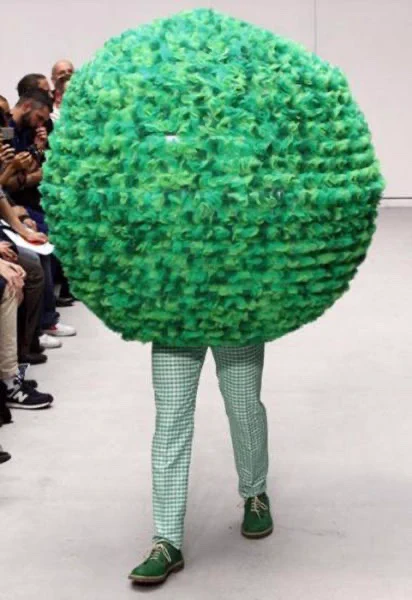 a fuzzy green ball wearing green pants strolls down the catwalk at a fashion show 