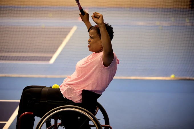 A woman in a wheelchair plays tennis and raises her arms in celebration