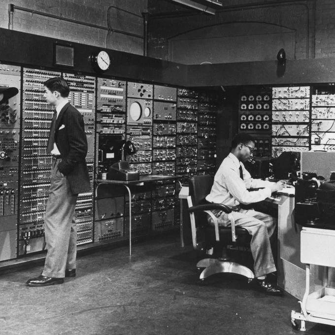 Black-and-white photo of Joe Thompson, seated, and Jack Gilmore, standing, work with a large room-sized computer