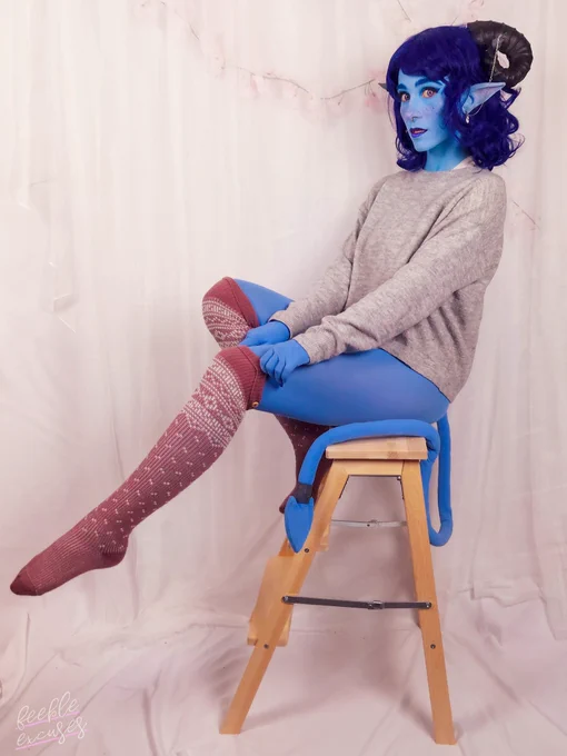 Photo of me as Jester from Critical Role. I am wearing a dark blue shoulder length wavy wig, black ram horns, long pointed ears and a blue tail.

All my skin is blue and I am wearing heavy makeup with a lot of pink blush and blue lipstick and eyebrows. I am wearing a light grey jumper and over the knee pink socks.

I am seating on a natural wood step stool, sideways, looking to the camera and holding up the sock on the leg closer to the camera, which is pointed forward.
