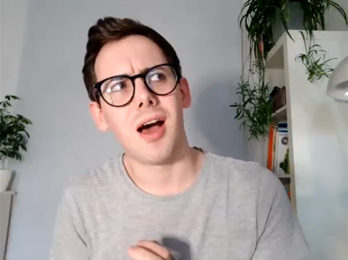 Zoom screen grab of Sab from Drag Queen Story Hour having a thoughtful and expressive moment. He is a young white man in a grey t-shirt with short dark hair and thick rimmed glasses. There are various plants behind him.