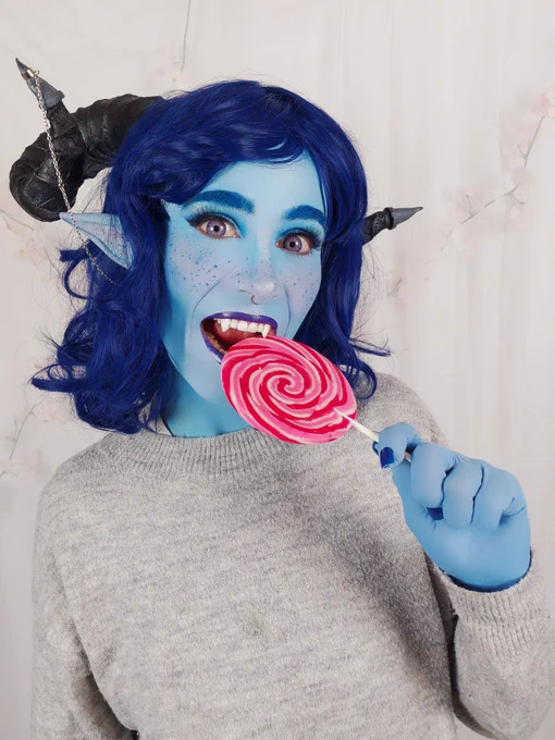 Photo of me dressed as Jester from Critical Role. I'm wearing a dark blue wig, black ram horns and long pointy ears. My skin is painted blue and I'm bitting on a pink lollipop