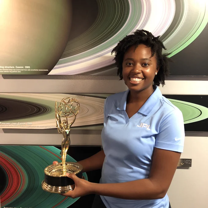 woman holding an emmy statuette while standing in front of a post with Cassini images of Saturn's rings