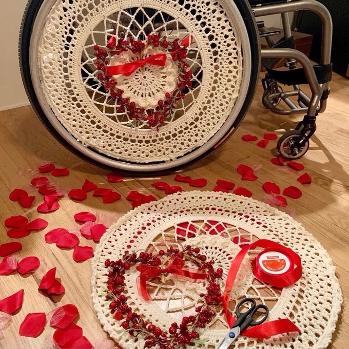 This image is the same as the first but Katie has the second, her left ivory wheel cover on the floor and she is working on decorating with another heart-shaped faux branch in its red faux berries, roll of red ribbon and a pair of scissor on top of the wheel cover.