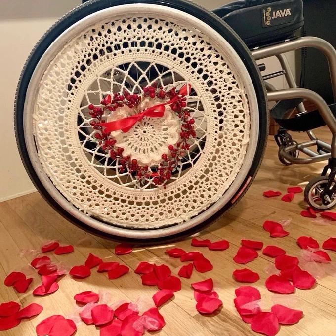 Side of Kate’s manual wheelchair with out beautiful crocheted ivory wheel cover. She has decorated with heart-shaped faux branch with red faux berries. She also attached red bow. She has red rose petals on the ground to set the vibe.