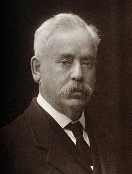 Picture of Frederick Mott in the early 20th century - an older man with white hair and a white moustache, on a dark background, wearing a suit, and looking somehow a bit lost.
