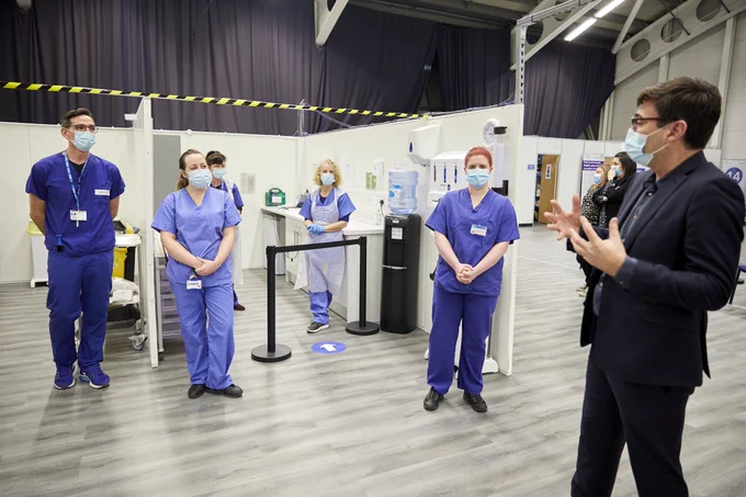 An image showing vaccination booths inside the Greater Manchester NHS Vaccination Centre, with Andy Burnham speaking to staff – all socially distanced and wearing masks.