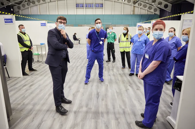 An image showing vaccination booths inside the Greater Manchester NHS Vaccination Centre, with Andy Burnham speaking to staff – all socially distanced and wearing masks.