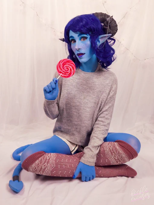 Photo of me dressed as Jester from Critical Role. I'm wearing a dark blue shoulder length wavy wig, black ram horns and long elf ears. My skin is painted blue and I'm wearing heavy pink blush and blue freckles. I'm wearing a light grey jumper and pink over the knee socks.