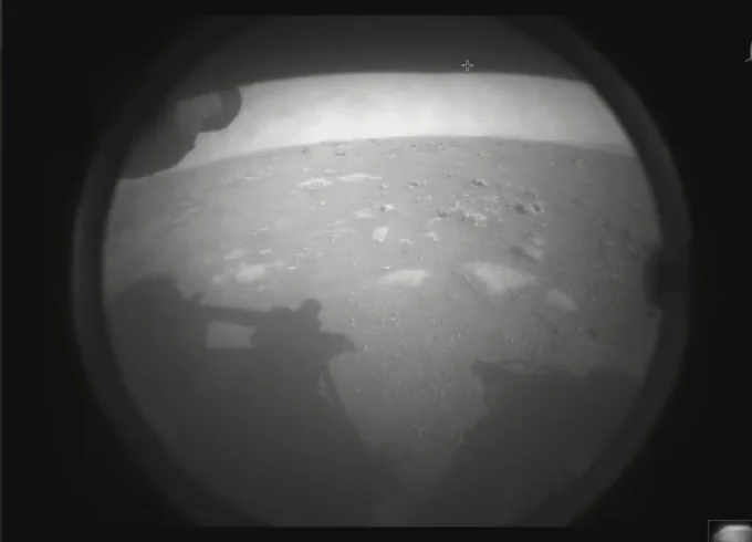 The first image taken by the Perseverance landing rover after a successful landing in Jezero Crater. The image is in grayscale. A circular lens, covered in a light film of dust, shows the Martian surface. Clusters of rocks can be seen scattered throughout the landscape.