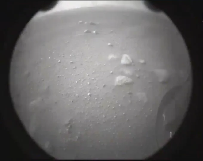 The second image taken by the Perseverance landing rover after a successful landing in Jezero Crater. The image is "behind" the lander. The image is in grayscale. A circular lens, covered in a light film of dust, shows the Martian surface. Clusters of rocks can be seen scattered in a pattern sweeping from the right to the upper left of the image.