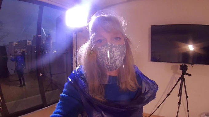A woman stares directly at the camera. She is wearing a blue snowflake mask and has shoulder length blonde hair. She is wearing a dark blue protective outfit with the hood at front. In the background is a camera on a tripod, in front of a large screen television against a white wall. In the  background is also a large glass door which we can see the back of the woman's reflection in with the stage light shining in it