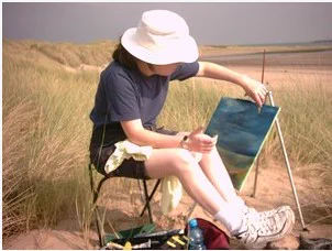 Photo of someone - perhaps CS Cape as a child - sat on a chair in the sand dunes, wearing black shorts, white trainers, blue t-shirt, shoulder length dark hair, white hat, with an easel next to them with a painting of the landscape - yellow sand, blue sea and sky