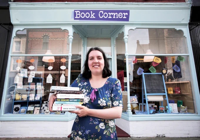 Photo of Jemma Warren in a patterned summery dress, shoulder length dark hair, big smile, looking straight at camera holding a stack full of books (The New Mrs Clifton by Elizabeth Buchan, The Curious Incident of the Dog in the Night by Mark Haddon, The Humans by Matt Haig, Chance Developments by Alexander McCall Smith, Jonathan Unleashed by Meg Rosoff and Us by David Nicholls) stood in front of her fantastic independent book shop, Book Corner, in Saltburn.