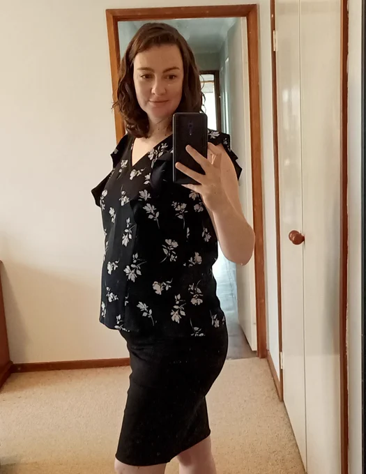 Kay is 6 months pregnant with a big belly. She wears a dark navy loose fitting top with small widely spaced white flowers, and a black pencil skirt, the image is cropped at the knee