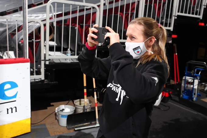 Kendall Coyne Schofield stands at ice level of the United Centre in Chicago, Ill. taking a photo of the arena with her phone. She wears a Chicago Red Stars face mask and a PWHPA hoodie. 