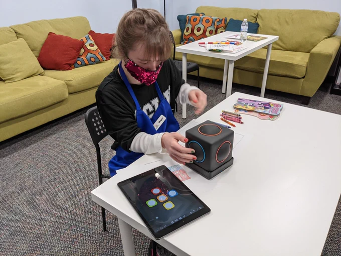 Young girl sitting at a table playing with a soft-sided black cube that color coordinates to sound (Skoog device).