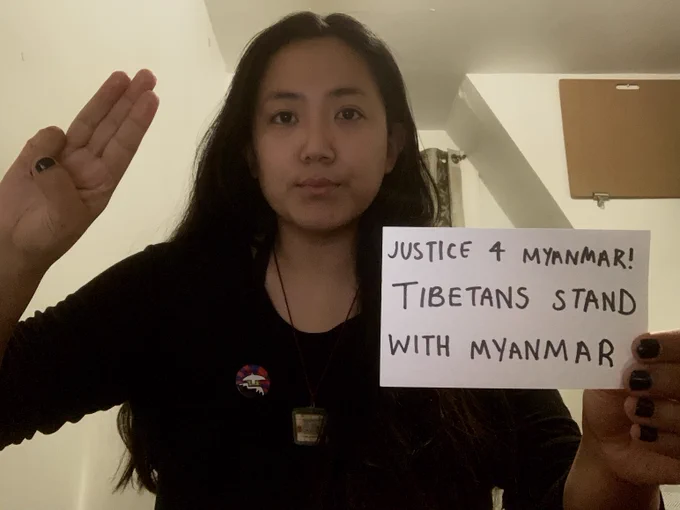 A Tibetan woman raising a three-finger salute in solidarity with the people of Myanmar, holding an index card with the text "Justice 4 Myanmar! Tibetans stand with Myanmar" and wearing a pin on her shirt of a white peace bird over the mountains of Tibet. 