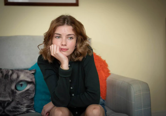 Teenage girl with brown hair sitting on sofa resting her chin in her hand. Girl is in contemplative mood