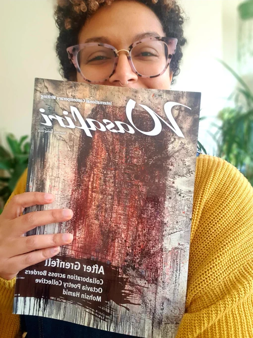 A woman wearing pink glasses and a yellow jumper holds up an issue of Wasafiri magazine. There are green plants in the background.