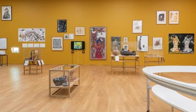 A view of MIMA’s collection gallery showing an array of artworks displayed on a golden yellow wall with a self-portrait by Sonia Boyce at the centre and cases of ceramics and jewellery in the foreground