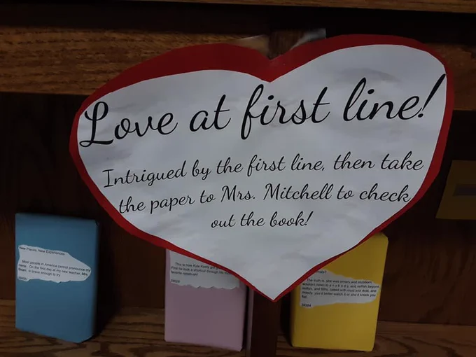 A red and white paper sign in the shape of a heart with the words, "Love at first line! Intrigued by the first line, then take the paper to Mrs. Mitchell to check out the book!"
