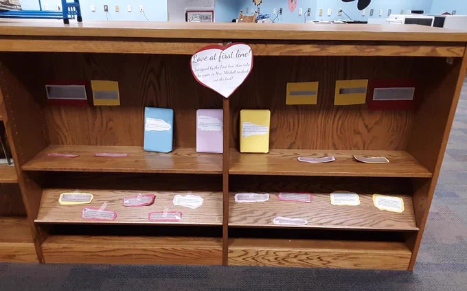 Books wrapped in paper to hide the cover are on a shelf. On each book is text that tells the first line of the book. A sign in the shape of a heart directs students to choose a book based on the quote for check out.