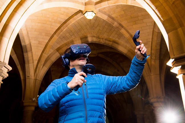 Dr Neil McDonnell using a VR headset, headphones and handheld controllers in the cloisters at the University of Glasgow's Gilmorehill campus.
