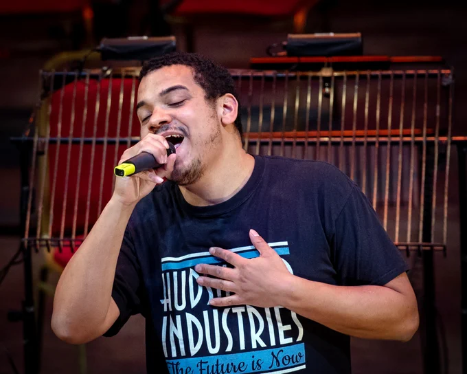 A person stands holding a microphone in this hand. They have their eyes closed, are wearing a blue t-shirt. Music stands are in the background. 