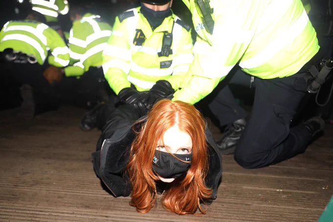 A woman on the ground wearing a black face mask with three police men on top of her holding her arms behind her back as they subdue her