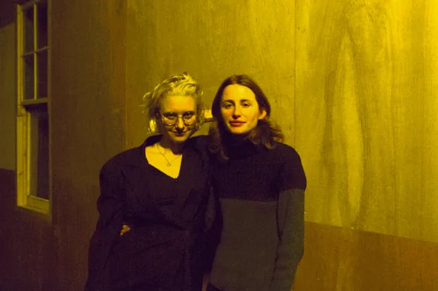 Two people, one with their arm around the other's waist, look at the camera. The woman on the right is smiling. Behind them is a hardboard wall with a window in the far left of the frame.