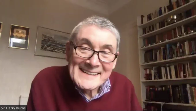 Harry Burns, wearing a red jumper smiles directly at the camera. Behind him are shelves of books 