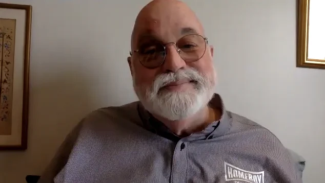 Father Gregory Boyle smiles into the camera. He is wearing a purple shirt with a logo that says 'Homeboy Industries'