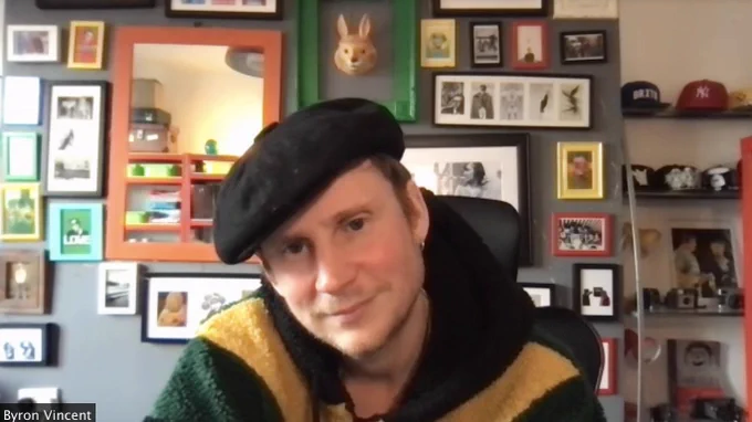 Byron smiles directly into the camera. He is wearing a black beret and behind him are numerous picture frames of varying sizes.