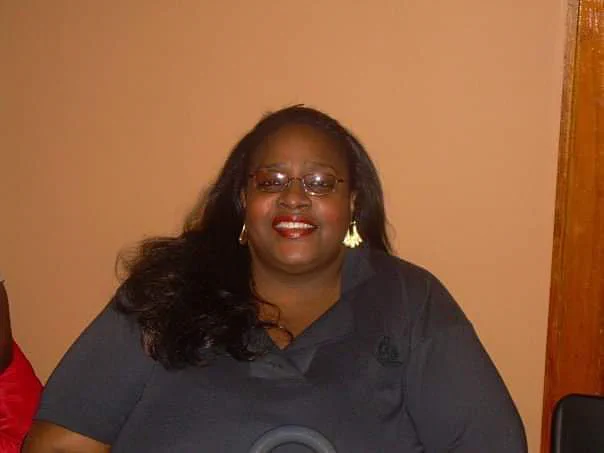 Photo of Patricia Frieson, an African American, retired nurse from Chicago who was the first COVID-19 victim in Cook County. In this photo she is wearing a gray polo, has long hair, has eyeglasses on and red lipstick. 