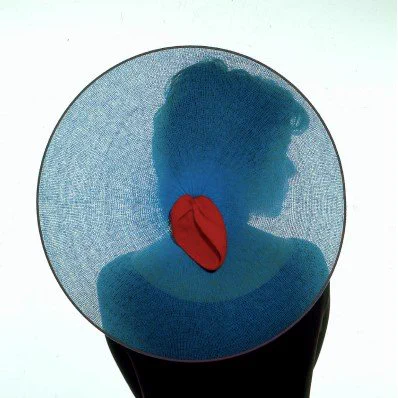 A female figure stands silhouetted from behind waist up. Against the back of her head is a netted circular blue headpiece with a piece of red cloth at its centre