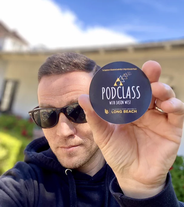 Host of Podclass with Jason West holds up a circular black sticker with the show’s name and logo.