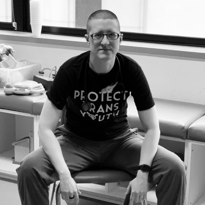 Androgynous white person wearing black rimmed glasses sitting in a chair looking seriously at the camera.  They wear a stethoscope and a black shirt which reads Protect Trans Youth.