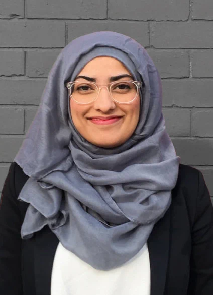A person wearing clear glasses and a grey hijab smiles directly at the camera while standing against a brick wall painted grey