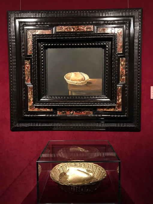 In an ornate black frame against a deep red wall, there is a painting of half a loaf of bread in a woven basket on the edge of a wooden table. In front of the painting on a plinth is what appears to be the bread depicted in the painting, only it and the basket have been painted gold.