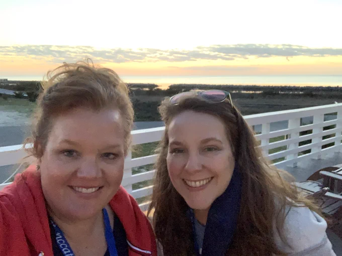 A photo of two women with a white fence, the NC coast, and a sunset behind them.