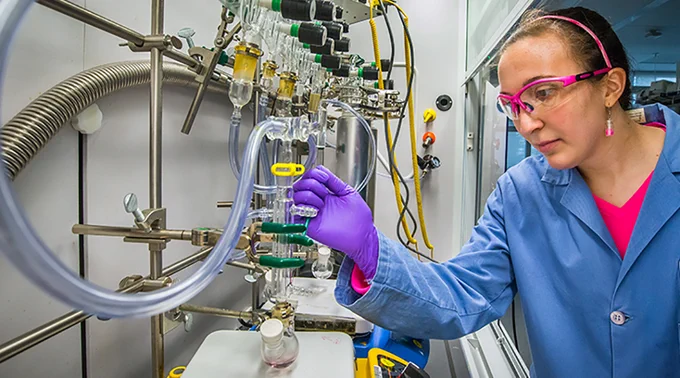 Photograph of a woman in lab goggles and a lab coat working with scientific equipment