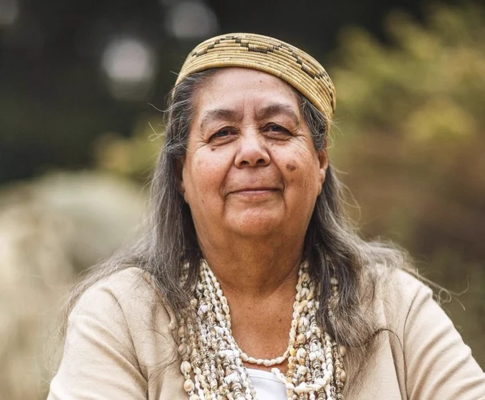 A portrait from the shoulders up of a person with long salt and pepper hair, wispy in the wind. They wear many necklaces made from sea shells. Their skin and shirt, soft earth tones. On their head is a sort of cap weaved from natural fibers with an intricate pattern. There face wears a subtle grin, they look calm and happy with eyes looking straight into the lens. A blurred background is composed of shrubs.