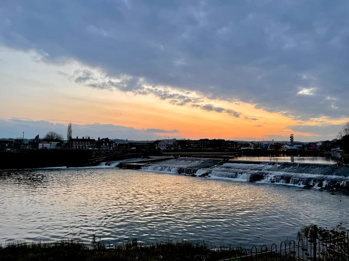 Trews Weir at sunset, looking up river. The sky is orange where clear but otherwise cloudy.