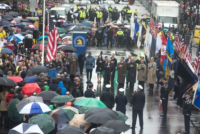 Photo from 2014: a rainy day in Boston where residents, first responders, and government officials -- including Secretary Marty Walsh and President Joe Biden-- stand in silence with their hands over their hearts.