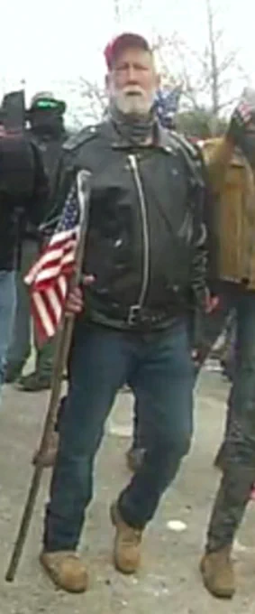 Capitol rioter 290 holding a American flag on a stick, wearing a black leather jacket, and wearing a red Trump hat.