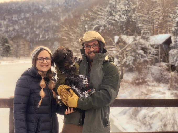 A woman and a man hold a puppy covered in snow in front of a winter landscape.